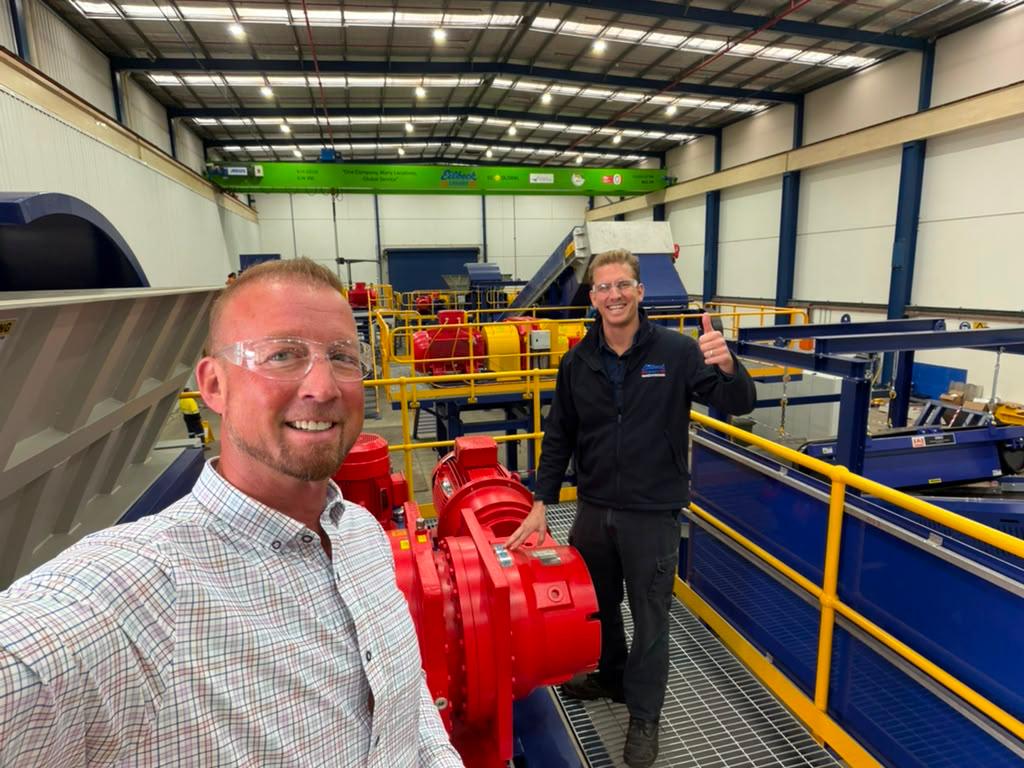 Two people smile for a selfie beside the red drives on the walkway of the shredding line