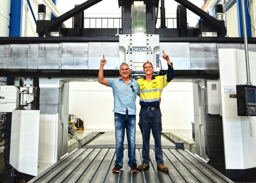 Two Eilbeck staff stand on the bed of a large gantry machining centre and point up at it